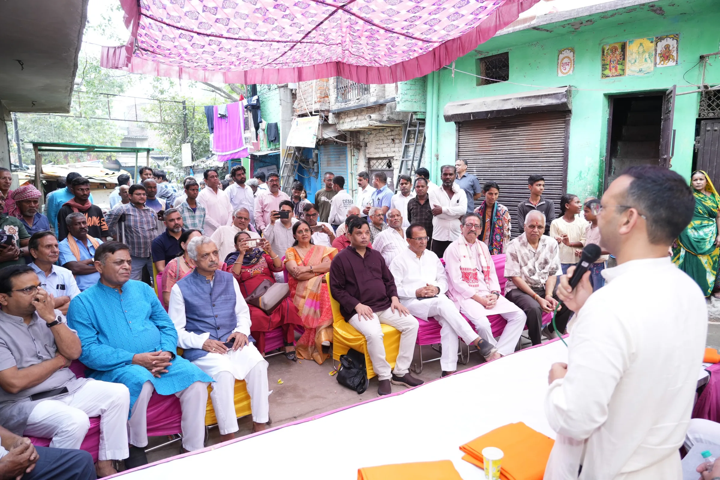 A large audience seated under a canopy in a lane, listening to a speaker with a microphone.